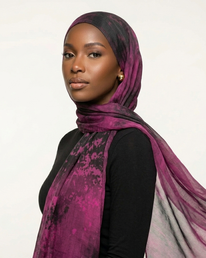 A woman wearing a sheer, lightweight hijab in a purple and black watercolor-like pattern, styled over a black top against a plain light background.