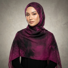A woman wearing a dark plum purple hijab with a black tie-dye or watercolor pattern, styled over a black top against a textured gray studio background.