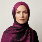 A woman wearing a draped hijab scarf in deep magenta and black with a mottled, watercolor-like pattern, set against a plain light gray background.