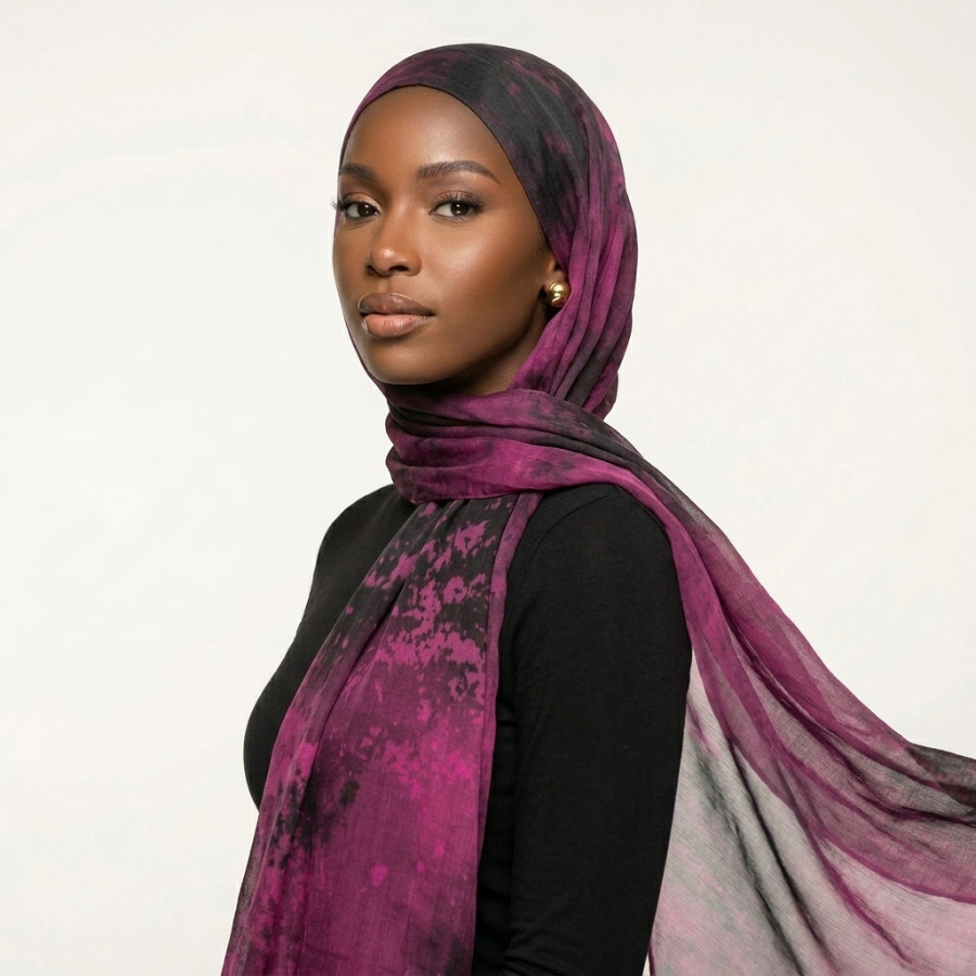 A woman wearing a sheer, lightweight hijab in a purple and black watercolor-like pattern, styled over a black top against a plain light background.