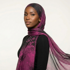 A woman wearing a sheer, lightweight hijab in a purple and black watercolor-like pattern, styled over a black top against a plain light background.